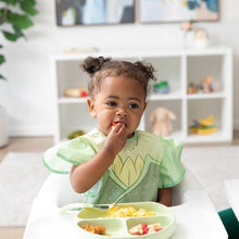 Load image into Gallery viewer, A toddler in a Bumkins Ruffle Apron Bib: Tiana eats a berry in a high chair, looking thoughtful, with toys on shelves behind.
