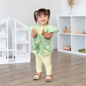 A young girl stands in a bright playroom, clapping her hands while wearing a Bumkins Ruffle Apron Bib: Tiana.