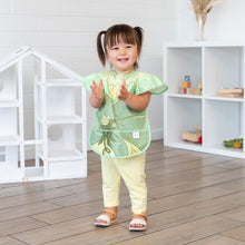 Load image into Gallery viewer, A young girl stands in a bright playroom, clapping her hands while wearing a Bumkins Ruffle Apron Bib: Tiana.
