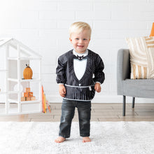 Load image into Gallery viewer, A boy wearing Bumkins Sleeved Bib: Jack Skellington stands in a bright playroom in front of a dollhouse, books, and gray chair.
