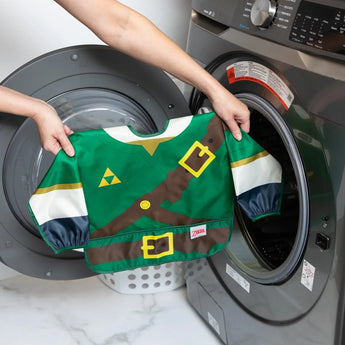 A person holds a Bumkins Sleeved Bib: Zelda™ in front of an open front-loading washing machine.