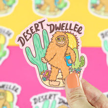 Load image into Gallery viewer, Orange-nailed hand holds Turtle Soups Desert Dweller Big Foot vinyl sticker: a scratch-resistant sasquatch with cactus and bottle.
