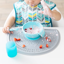 Load image into Gallery viewer, A baby messily eats yogurt and strawberries from a Bumkins Silicone First Feeding Set: Blue on a gray placemat with a blue cup nearby.
