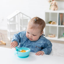 Load image into Gallery viewer, A toddler in a Bumkins Sleeved Bib: Vintage Denim eats from a blue bowl with a matching spoon at a white table in a bright playroom.
