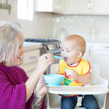 Load image into Gallery viewer, In a bright kitchen, a woman feeds a baby in a high chair wearing a yellow bib from a Bumkins Silicone First Feeding Set: Blue.
