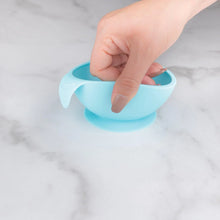 Load image into Gallery viewer, A hand holds a Bumkins Silicone First Feeding Set: Blue, demonstrating the suction cup bottom on a marble surface.
