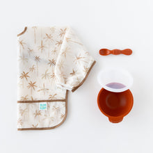 Load image into Gallery viewer, Bumkins Silicone First Feeding Set: Clay rests next to a folded beige &amp; brown palm tree print bib on a white background.
