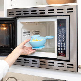 A hand places a Bumkins Silicone First Feeding Set: Mickey Mouse Light Blue filled with food into an open microwave.