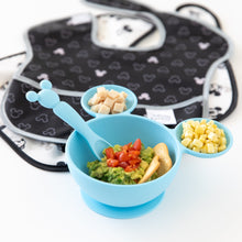 Load image into Gallery viewer, Bumkins Silicone First Feeding Set: Mickey Mouse Light Blue, with mashed avocado and corn, sits next to stacked bibs on a white background.
