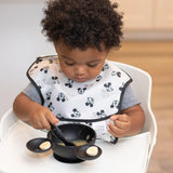 A toddler in a high chair enjoys bananas and applesauce from a Bumkins Silicone First Feeding Set: Mickey Mouse Classic Black.