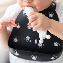 Load image into Gallery viewer, A child chews a spoon from Bumkins Silicone First Feeding Set: Mickey Mouse Vanilla Sprinkle while wearing a black silicone bib.
