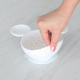 A hand lifts a Bumkins Silicone First Feeding Set: Mickey Mouse Vanilla Sprinkle on a marble surface, showcasing the suction cup bottom.