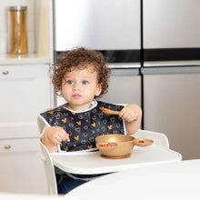 Load image into Gallery viewer, A toddler with curly hair eats from a Bumkins Silicone First Feeding Set: Minnie Mouse Gold in a modern kitchen while wearing a black bib.
