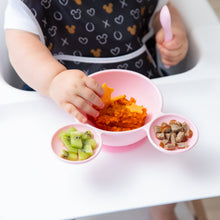 Load image into Gallery viewer, A toddler in a highchair eagerly explores the a Bumkins Silicone First Feeding Set: Minnie Mouse Light Pink, reaching for mashed sweet potato.
