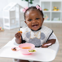 Load image into Gallery viewer, A happy baby sits in a highchair using the Bumkins Silicone First Feeding Set: Minnie Mouse Light Pink while wearing a matching bib.
