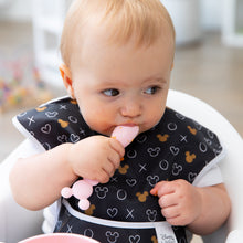 Load image into Gallery viewer, A baby with a patterned bib curiously chews a spoon from Bumkins Silicone First Feeding Set: Minnie Mouse Light Pink, looking aside.
