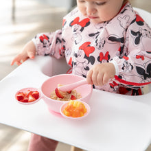Load image into Gallery viewer, A baby in a bib self-feeds oatmeal and strawberries from Bumkins Silicone First Feeding Set: Minnie Mouse Light Pink at a white highchair.
