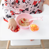 A child enjoys oatmeal with fruit from Bumkins Silicone First Feeding Set: Minnie Mouse Light Pink on their high chair tray.