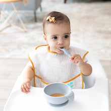 Load image into Gallery viewer, A baby with a bow and beige bib sits in a high chair, enjoying puree from a Bumkins Silicone First Feeding Set: Gray.
