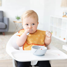 Load image into Gallery viewer, A baby wearing a yellow bib in a high chair eats from a Bumkins Silicone First Feeding Set: Gray, with a chair and shelves in the background.
