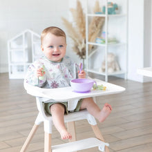 Load image into Gallery viewer, A baby in a high chair wearing a floral bib eats from a Bumkins Silicone First Feeding Set: Lavender with green utensils in a bright room.
