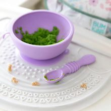 Load image into Gallery viewer, Close-up of Bumkins Silicone First Feeding Set: Lavender filled with green mashed food on a white silicone mat adorned with food crumbs.
