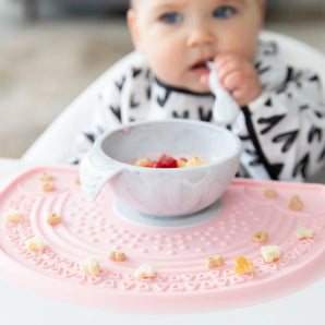 A baby in a black & white bib eats from a Bumkins Silicone First Feeding Set: Marble on a pink silicone mat covered in cereal pieces.