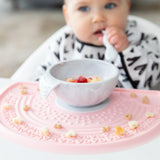 A baby in a black & white bib eats from a Bumkins Silicone First Feeding Set: Marble on a pink silicone mat covered in cereal pieces.