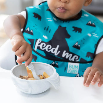 A child eagerly eats oatmeal from a Bumkins Silicone First Feeding Set: Marble, wearing a blue bear-patterned bib in a high chair.