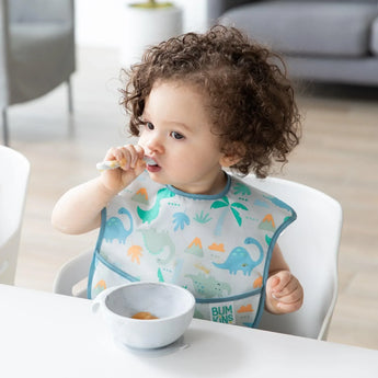 A curly-haired toddler in dinosaur bib eats from a Bumkins Silicone First Feeding Set: Marble at a white table.