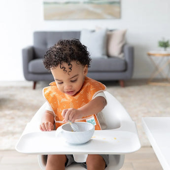 A toddler with curly hair sits in a high chair, wearing an orange bib and eating from a Bumkins Silicone First Feeding Set: Marble.