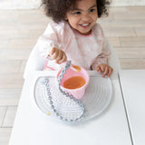 A curly-haired child in a pink bib beams at a white table, eating from a Bumkins Silicone First Feeding Set: Pink on a gray placemat.