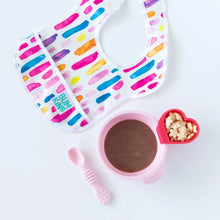 Load image into Gallery viewer, A colorful bib sits next to Bumkins Silicone First Feeding Set: Pink with puree and a red heart-shaped cup of cereal on a white background.
