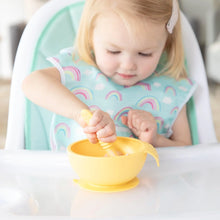 Load image into Gallery viewer, A child in a high chair wearing a rainbow-print bib uses a yellow Bumkins Silicone Dipping Spoons 3 Pack: Tutti-Frutti to eat from a bowl.
