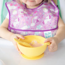 Load image into Gallery viewer, A child in a high chair in a purple bib holds a yellow bowl with a yellow Bumkins Silicone Dipping Spoons 3 Pack: Tutti-Frutti inside.
