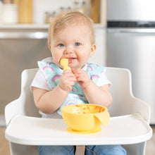 Load image into Gallery viewer, A baby wearing a rainbow-print bib in a high chair self-feeds from a Bumkins Silicone First Feeding Set: Pineapple.

