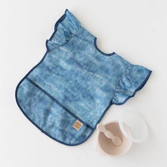 Bumkins Ruffle Apron Bib: Crushed Denim lays flat on a white background next to a beige bowl & spoon set.