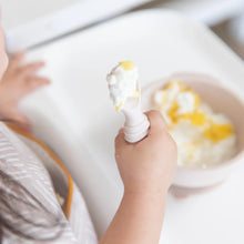 Load image into Gallery viewer, Close-up of a child holding a spoon from Bumkins Silicone First Feeding Set: Sand covered in yogurt, with the bowl blurred underneath.
