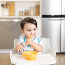 Load image into Gallery viewer, A toddler wearing a white and blue bib in a high chair eats from a Bumkins Silicone First Feeding Set: Tangerine in a kitchen.
