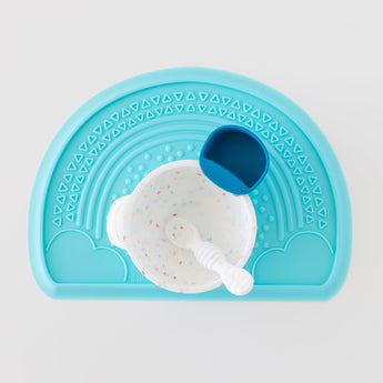 Bumkins Silicone First Feeding Set: Vanilla Sprinkle sits on top of a blue placemat with a dark blue cup on a white background.