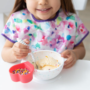 A child in a floral bib beams while enjoying ice cream from a Bumkins Silicone First Feeding Set: Vanilla Sprinkle  at a white table.