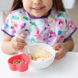 A child in a floral bib beams while enjoying ice cream from a Bumkins Silicone First Feeding Set: Vanilla Sprinkle  at a white table.