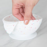 A hand holds a Bumkins Silicone First Feeding Set: Vanilla Sprinkle, demonstrating the suction cup bottom on a marble surface.