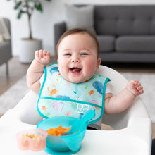 Load image into Gallery viewer, A smiley baby wearing a Bumkins Starter Bib 2 Pack: Ocean Life &amp; Whale Tail sits in a high chair with a blue bowl of mashed food on the tray.
