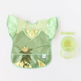 Bumkins Ruffle Apron Bib: Tiana lays next to a green bowl, spoon, & lid set on a white background.
