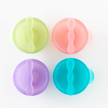 Top down view of 4 colorful bowls with spoons, including Bumkins Silicone First Feeding Set: Green Jelly, neatly arranged on a white background.