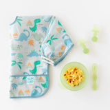 Bumkins Silicone First Feeding Set: Green Jelly with eggs sits next to a folded dinosaur bib & green toddler utensils on a white background.