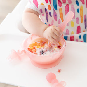 Close-up of a baby in a colorful bib eating yogurt & fruit from a Bumkins Silicone First Feeding Set: Pink Jelly near pink toddler utensils.