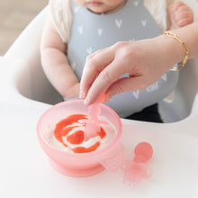 Load image into Gallery viewer, Close-up of an adult hand dipping a spoon into yogurt in a Bumkins Silicone First Feeding Set: Pink Jelly in front of a baby in a gray bib.
