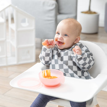 A smiling baby in a gray & white bib messily eats from a Bumkins Silicone First Feeding Set: Pink Jelly while sitting in a high chair.